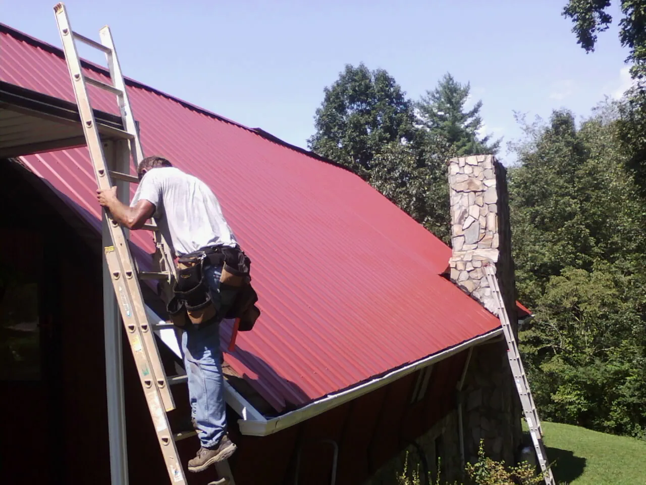 Roofer on a ladder beside a red metal roof and stone chimney in Milton